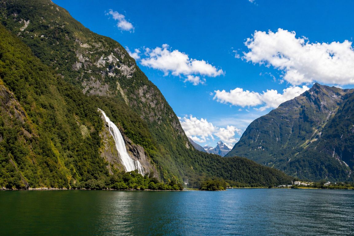 Lady Bowen Falls waterfall in Fiordland National Park with dramatic alpine peaks and blue skies, seen during a guided trip with New Zealand Milford Sound Tours